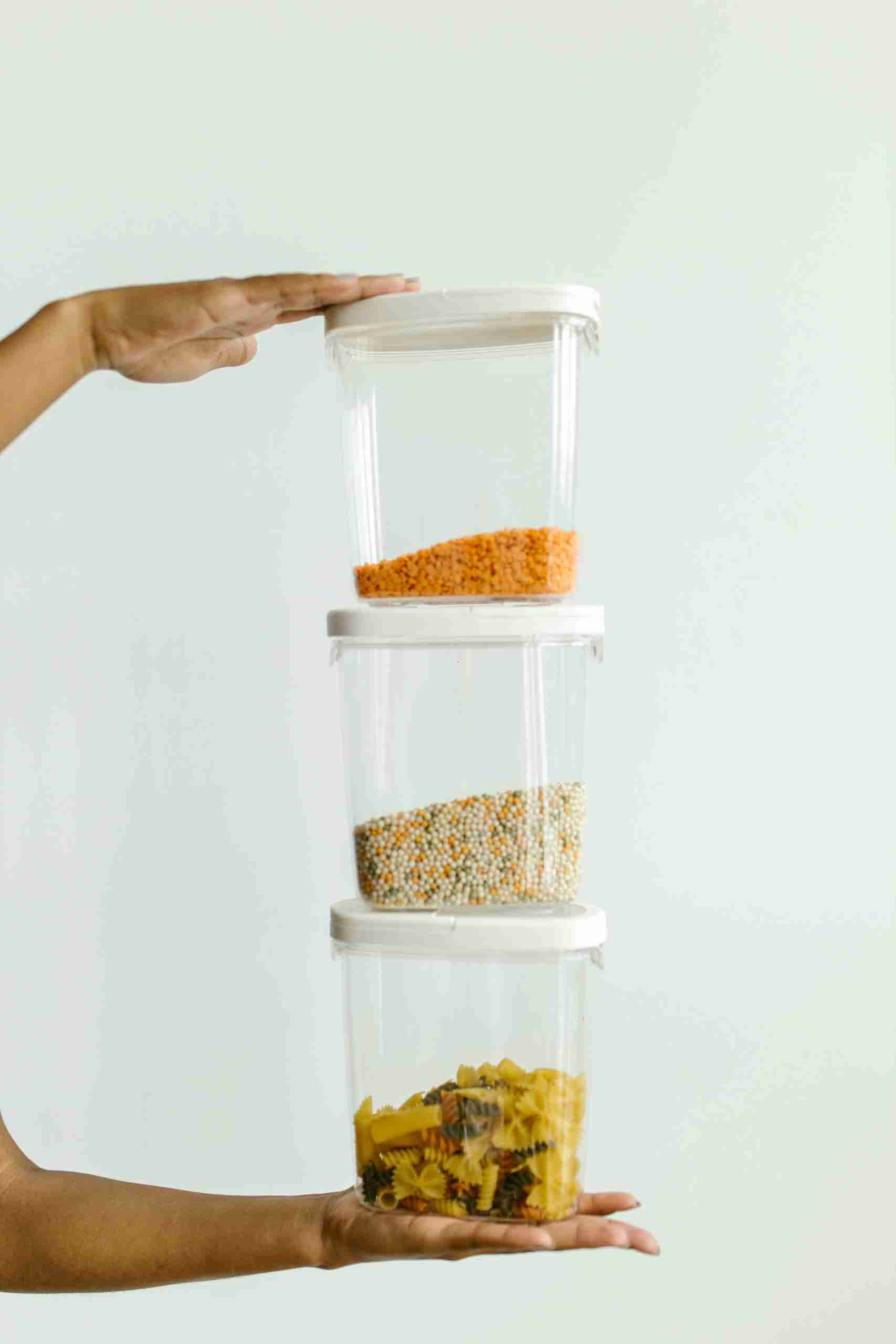 Hands holding stacked plastic containers with various dry foods against a neutral background.